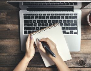 klawiatura laptopa i leżący na niej notatnik oraz długopis, oba trzymane przez kobiecie dłonie